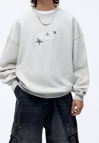 Persona che indossa un ampio sweatshirt grigio chiaro con testo e stelle, pantaloni cargo neri, una collana in argento e anelli alle dita.