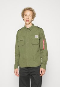 Olive green button-up shirt, chest pockets, patch on left side, and a red zipper pull accent on right sleeve. Cotton material, relaxed fit.