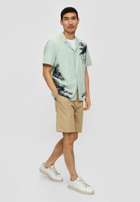 Lichtgroen overhemd met korte mouwen en zwarte palmbomenprint, gecombineerd met beige shorts en witte sneakers. Eenvoudig ontwerp met knoopsluiting.