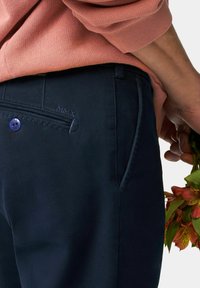 Les chinos navy présentent une coupe slim, des poches avant, une poche arrière avec un bouton visible et des coutures. Un logo MMX discret est embouti sur la poche.