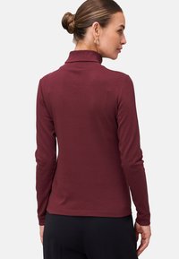 Woman with hair in a bun wearing gold hoop earrings, a long-sleeve burgundy turtleneck sweater, and black pants, facing away.
