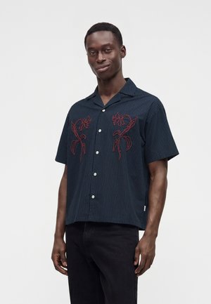 Homme portant une chemise bleu foncé à manches courtes avec de fines rayures et une broderie florale rouge sur la poitrine, associée à un pantalon noir.