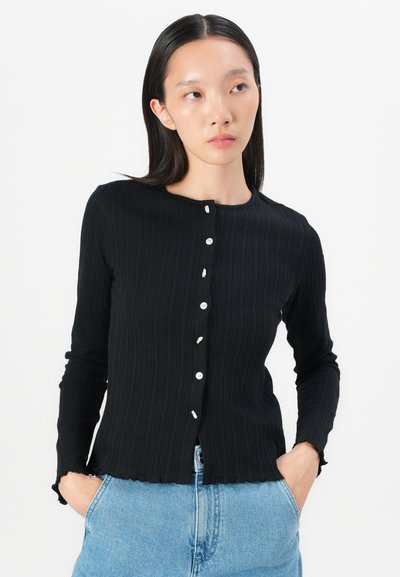 Black ribbed long-sleeve shirt with a rounded neckline, front button closure, scalloped hem, and textured fabric. Paired with light blue jeans.