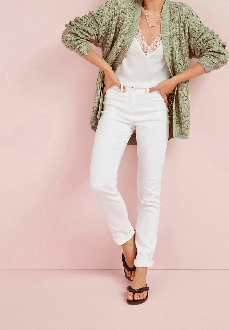 Hellgrüne gestrickte Strickjacke, weißes Top mit Spitzenbesatz, weiße eng anliegende Jeans, braune Flip-Flops, vor einem pinken Hintergrund präsentiert.