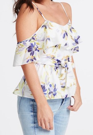 Kobieta w białym bluzce typu off-shoulder w kwiatki fioletowe i żółte, z falbankami, przewiązana w pasie i w jasnoniebieskich jeansach.