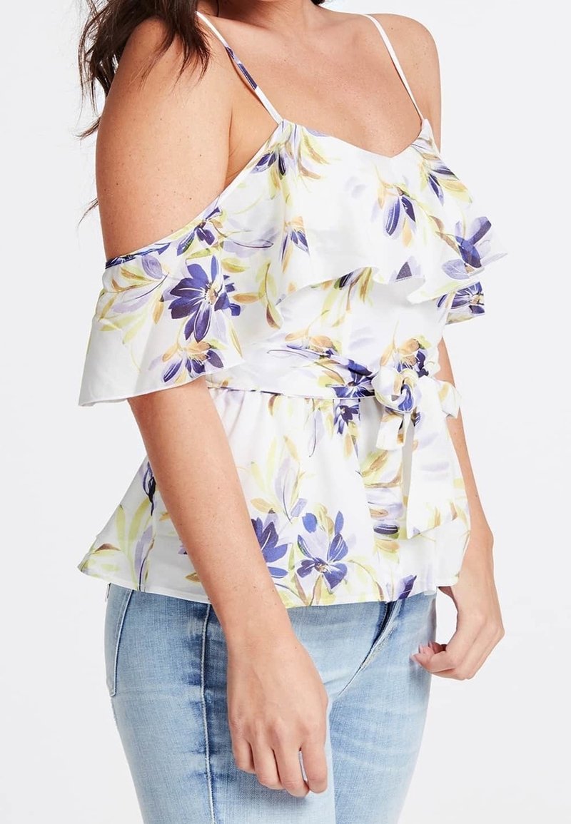 Femme portant un haut blanc à épaules dénudées avec des fleurs violettes et jaunes, détails à volants, taille nouée, et jean bleu clair.