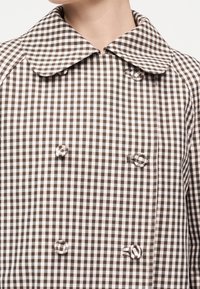 Καφέ και λευκό καρό διπλό παλτό με μεγάλο στρογγυλεμένο γιακά και κουμπιά καλυμμένα με ύφασμα, φορεμένο από άτομο με εμφανή λαιμό και πηγούνι.