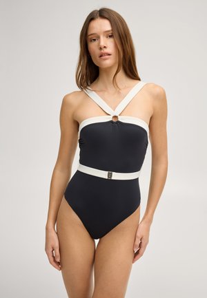 Femme portant un maillot de bain une pièce noir avec des bretelles blanches croisées sur la poitrine et une ceinture blanche à la taille, debout devant un fond gris.