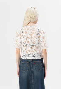 Blouse blanche texturée avec des manches ballon, ornée d'un motif floral en dentelle complexe. Portée sur une jupe en denim foncé avec des poches arrière visibles.