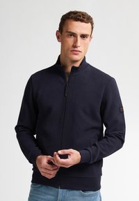 Granatowa bluza zapinana na zamek, wykonana z teksturowanej tkaniny w diamentowy wzór, z wysokim kołnierzem i ściągaczami przy mankietach. Prosty design z minimalnymi akcentami.