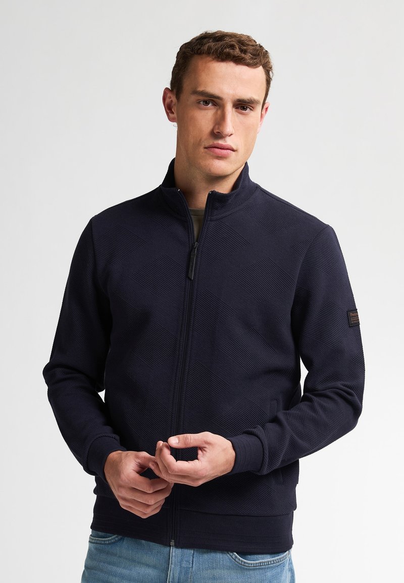 Granatowa bluza zapinana na zamek, wykonana z teksturowanej tkaniny w diamentowy wzór, z wysokim kołnierzem i ściągaczami przy mankietach. Prosty design z minimalnymi akcentami.