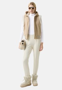 Beige Weste mit Kapuze und Schaffellbesatz, weißer Reißverschluss-Oberteil, cremefarbene Hose und beige, mit Fell gefütterte Stiefel; hält ein kleines Felltäschchen.