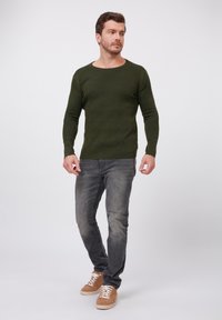 Suéter acanalado de color verde oliva con cuello redondo, combinado con jeans gris descolorido y zapatillas marrón claro con cordones blancos.