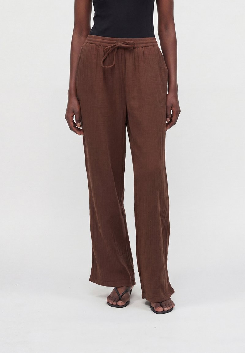 Personne portant un pantalon marron ample avec cordon, un top noir sans manches et des sandales noires, debout sur un fond blanc uni.
