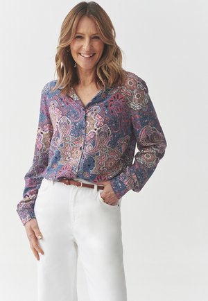 Long-sleeve button-up blouse featuring a multicolored paisley pattern on a navy background. Worn with white pants and a brown belt.