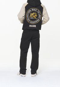 Felpa con cappuccio nera e beige con un logo sulla schiena, presenta un cappuccio regolabile, abbinata a pantaloni neri e sneakers bianche.