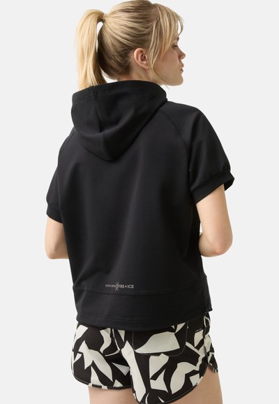Schwarzer kurzärmliger Hoodie mit einer taillierten Passform, ausgestattet mit einer Kapuze und weißen Logodetails am Saum. Kombiniert mit gemusterten Schwarz-Weiß-Shorts.