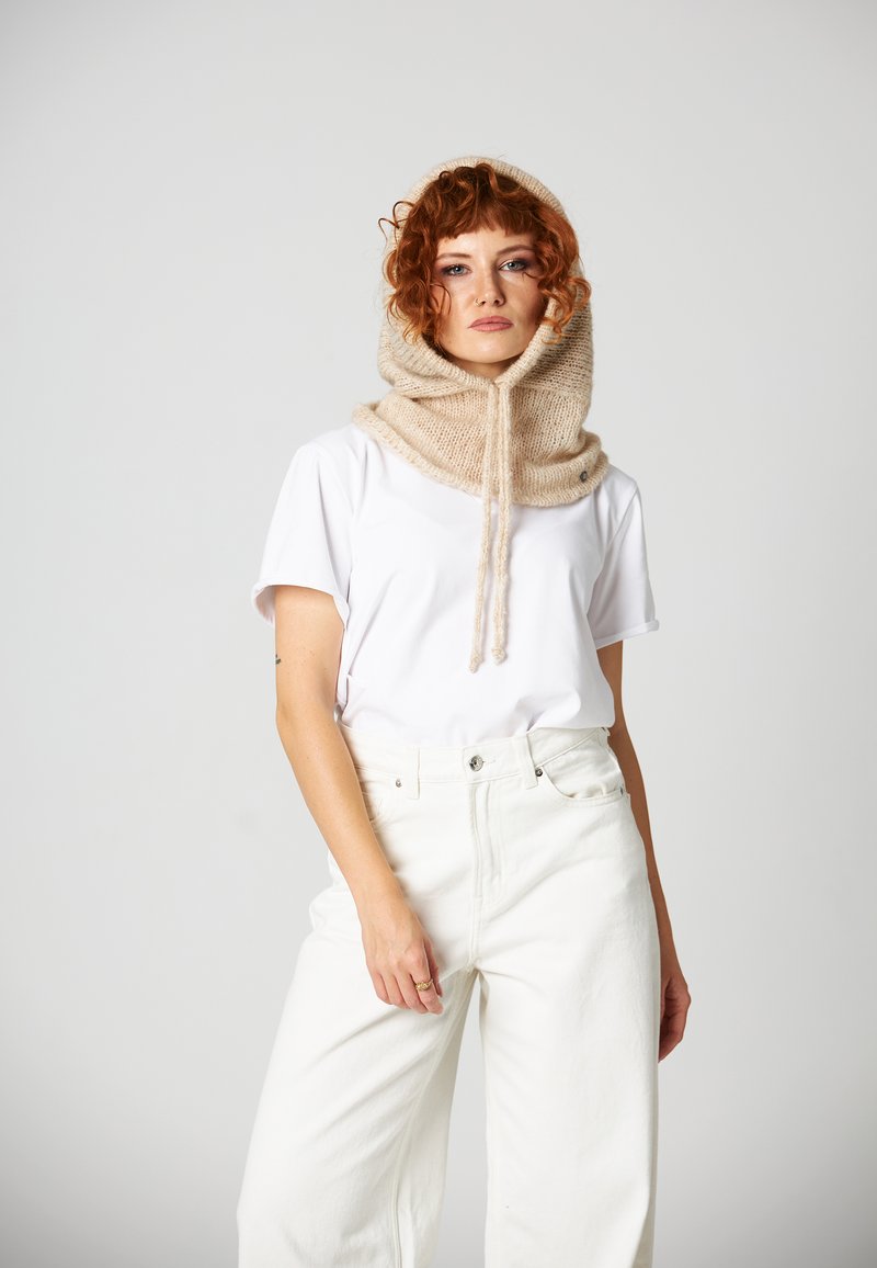 Beżowy, dziany szal-kaptur z teksturowanym wykończeniem, z regulowanymi sznurkami. Nosi się go na gładkiej, białej koszulce i białych spodniach.