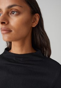 Schwarzes geripptes Rollkragenoberteil mit hohem Kragen, das eine glatte Textur und einen taillierten Schnitt aufweist, teilweise an einer Person sichtbar.