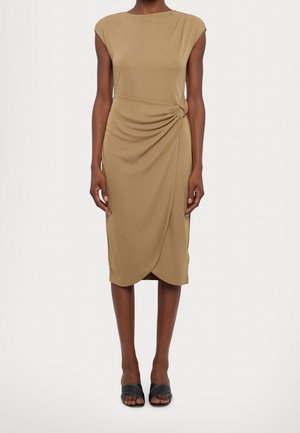 Femme portant une robe sans manches beige longueur genou avec un détail nœud sur le côté et des talons ouverts noirs, debout devant un fond uni.