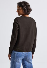 Jeune femme aux cheveux courts et bouclés, portant un haut à manches longues marron foncé et un jean en denim bleu clair, vue de dos et regardant sur le côté.