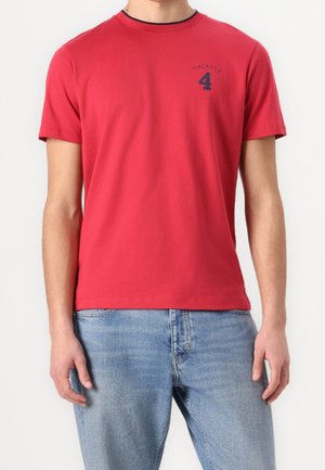 Mann trägt ein rotes kurzärmeliges T-Shirt mit „Hackett 4“ in Marineblau auf der Brust, kombiniert mit hellblauen Jeans.