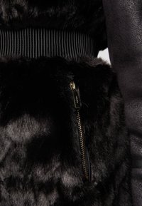 Czarna kurtka z sztucznego futra z teksturowanym materiałem, prążkowanym czarnym pasem w talii oraz złotymi detalami w postaci zamków błyskawicznych w kieszeniach.
