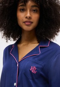 Γυναίκα με σγουρά μαλλιά φορώντας μπλε ναυτικό μπλουζάκι πιτζάμας με ροζ περιθώριο και κεντημένο λογότυπο «RLL» στην τσέπη στο στήθος.