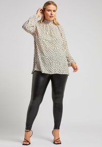Blusa beige a pois con maniche lunghe e collo alto abbinata a leggings neri lucidi e tacchi neri con cinturini.