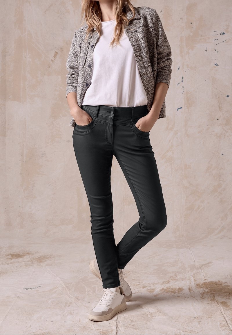 Femme portant un T-shirt blanc, une veste grise à motifs, un pantalon noir slim et des baskets blanches, debout les mains dans les poches contre un mur beige.