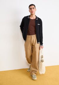 Giacca nera con zip e rifiniture bianche e dettagli ricamati, camicia marrone aderente, pantaloni a gamba larga color sabbia e borsa a tracolla beige trapuntata, abbinati a sneaker chiare.