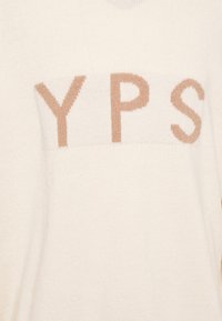 Pull en maille couleur crème avec une texture douce. Présente de grandes lettres "YPS" en marron clair sur le devant, créant un contraste simple.