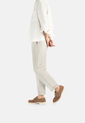 Camicia leggera color crema con taschino, abbinata a pantaloni grigi chiari e scarpe in suede marroni con suole bianche. Sottile trama nei materiali.