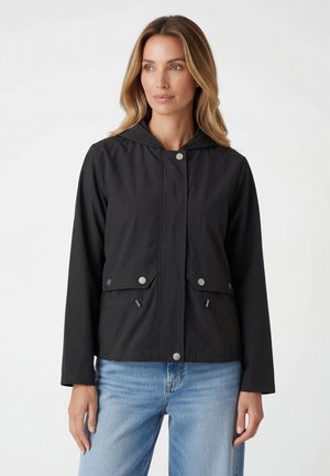 Frau trägt eine schwarze Kapuzenjacke mit silbernen Knöpfen und Reißverschlusstaschen, kombiniert mit hellblauen Jeans, stehend vor weißem Hintergrund.