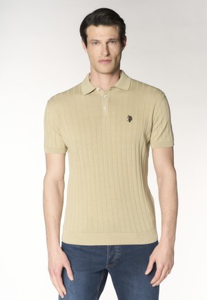 Uomo che indossa una polo beige a maniche corte con coste verticali e jeans blu, in piedi davanti a uno sfondo chiaro uniforme.