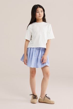 Niña de pie con cabello largo y negro, vistiendo una camiseta blanca de manga corta con detalles florales, falda azul a rayas y zapatillas beige.