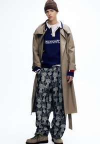 Bež kabast kaput, plavi pulover s "STWD" logotipom, tamne hlače s uzorkom koji prikazuje lubanje i stubove, smeđa kapa, bež cipele.