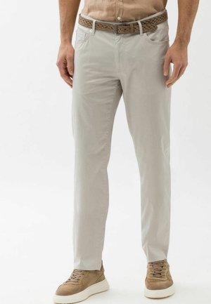 Homme portant un pantalon gris clair, une ceinture tressée marron, des baskets beige et une chemise beige boutonnée, debout devant un fond blanc uni.