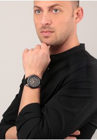 Orologio in gomma nera con cassa rotonda caratterizzato da accenti dorati e numeri arabi. Modello che indossa una camicia nera con colletto, mano appoggiata vicino al mento.