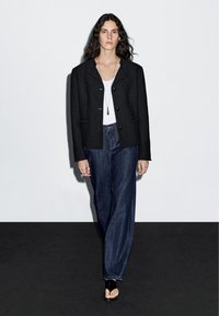 Žena s dlouhými tmavými vlasy, oblečená v černém saku, bílém topu, širokých džínách, černých otevřených botách a s vícevrstvými náhrdelníky, stojící na černé podlaze.