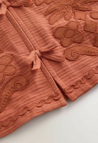 Nærbillede af rustfarvet ribbet stof med broderede blomsterprint og en bundet stofknude langs sømmen.