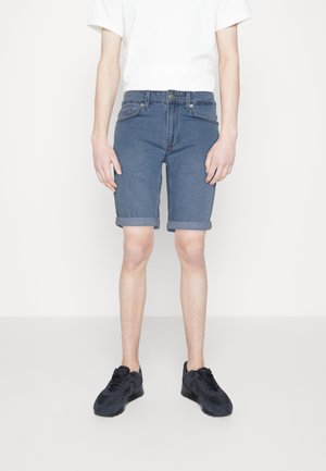 Pantalones cortos de mezclilla en azul, con un corte recto, dobladillo enrollado y diseño de cinco bolsillos. Combinados con zapatillas negras y una camiseta blanca.