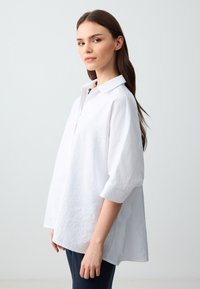 Giovane donna con lunghi capelli castani, indossa una camicia bianca sbottonata e pantaloni scuri, in piedi contro uno sfondo chiaro e semplice, rivolgendosi di lato.