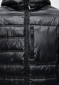 Veste matelassée noire brillante avec une fermeture éclair frontale et une poche poitrine zippée verticale sur le côté droit.