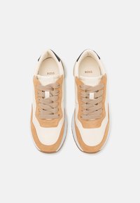 Beige und tanfarbene Sneakers aus Leder und Wildleder, mit einer glatten Textur, gepolsterter Zunge und flachen Schnürsenkeln. Der Markenname ist innen sichtbar.