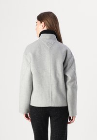 Grå uldjakke med høj krave og droppede skuldre. Har et syet mønster og prægede logo bagpå. Glat tekstur.