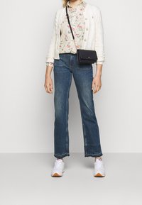 Weißer Kabelstrick-Cardigan über einem floral gemusterten Hemd, hoch taillierte blaue Jeans und weiße Sneakers mit orangefarbenen Akzenten. Kompakte schwarze Umhängetasche.
