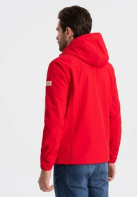 Giacca con cappuccio rossa realizzata in tessuto liscio, caratterizzata da cuciture laterali e un piccolo badge sulla manica sinistra. Indossata con jeans blu.