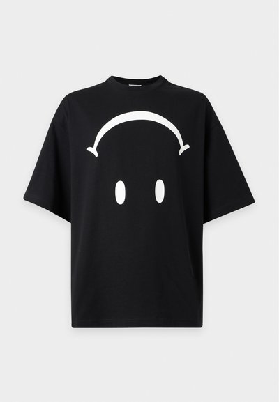 T-shirt noir à manches courtes ample avec un grand motif de visage minimaliste blanc comprenant deux yeux ovales et une ligne courbée au-dessus.