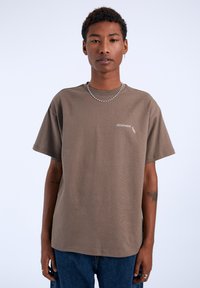 Camiseta de algodón marrón con mangas cortas, un escote redondo y un pequeño logo en el lado izquierdo del pecho. Combinada con jeans de mezclilla azul.